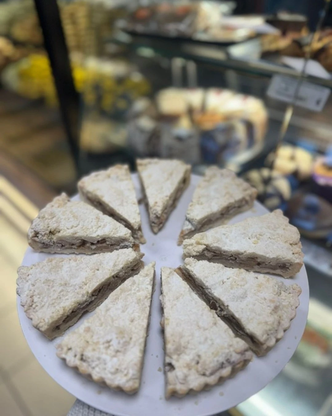 konak-bakery-stuttgart-apfelkuchen-puderzucker-2 Ein Teller mit frisch gebackenen, mit Puderzucker bestäubten Apfelkuchenstücken vor dem Schaufenster der Konak Bakery in Stuttgart.